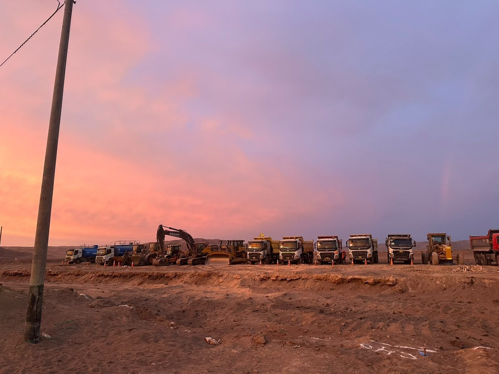 Maquinaria pesada y volquetes alineados al atardecer en un terreno desértico.