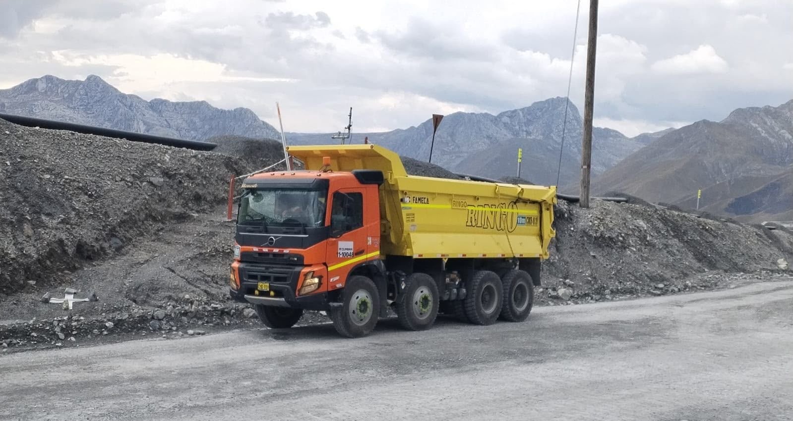 Volquete Ringo con cabina naranja y tolva amarilla en una vía minera, con cordillera al fondo.